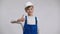 Joyful Caucasian boy showing thumb up posing at white background in hard hat and blue overalls. Portrait of happy