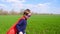 Joyful child in a superhero costume, a red cloak and a mask runs through the green grass, smiling and looking toward the