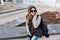 Joyful fashionable young woman in warm winter clothes, knitted hat, sunglasses sitting on street in city, speaking on