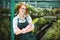 Joyful florist in apron and pink gloves standing with little garden shovel in hand. Young smiling lady in eyeglasses