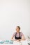 Joyful girl in black apron and striped T-shirt sitting at the table with handmade plates happily looking aside at