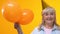 Joyful grandmother in party hat showing orange balloons on yellow background