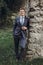 Joyful groom in elegant blue suit smiling and posing near old castle wall, handsome businessman posing outdoors, newlywed groom