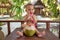 A joyful and happy little one year old child sits on a wooden table and drinks coconut milk from fresh green coconut through a