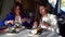 Joyful ladies are dining in restaurant at daytime, smiling and cutting meals, portrait of two female friends