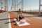 Joyful lady in stylish tracksuit sits by modern laptop on empty footbridge