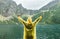 Joyful person in yellow raincoat with raised hands stands against unreal mountain landscape with lake. Person hiking on Lake