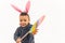 Joyful playful multiracial baby boy in pink fuzzy bunny ears hairband posing over white background with cardboard Easter