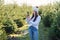 Joyful pretty young woman in warm hat photography beautiful christmas trees during walking among forestry plantings.