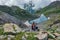 Joyful successful man tourist with victory gesture on rock at mountains near cold mountain lake