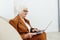 a joyful, sweet, gray-haired elderly woman in a brown suit is sitting on a beige sofa in a bright room working remotely