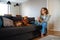 Joyful white woman using cellphone while sitting on sofa with her dog