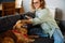 Joyful white woman using cellphone while sitting on sofa with her dog