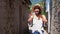 Joyful young girl tourist in white shirt and straw hat walking on ancient street