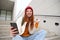 Joyful young redhead girl sits on street stairs, say yes, wins and celebrates with phone in hand, receive positive news