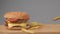 A juicy burger is lying on a wooden table and fries are falling down next to it.