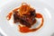 Juicy piece of lamb under red sweet sauce and covered with fried onion on large round white plate on white background.