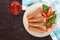 Juicy sausages with tomatoes and basil leaves on a plate on a dark wooden background.