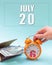 July 20th. Hand holding an orange alarm clock, a wallet with cash and a calendar date. Day 20 of month.