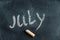July. Name of summer month written in yellow chalk on black chalkboard. Handwritten text. A piece of colored chalk lies next to it