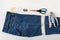 Just cutted blue denip shorts, measuring tape, scissors and sewing pin on a white table. Shorten the jeans with scissors and