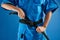 Karate fighter in blue kimono uniform tying black belt with hands close-up on blue background with copy space isolated in studio,