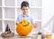 kid in apron painting Halloween pumpkin, at wooden table, preparing holiday decorations at home