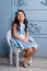 A kid in a blue dress, beige heels, sitting on a white chair, on a gray background.