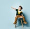 Kid in colorful t-shirt, brown pants. Smiling, pointing at something, sitting on white chair against blue background