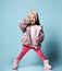 Kid with headband, in pink faux fur coat, pants and boots. She smiling, gesticulating, posing on blue background. Full length