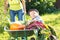 Kid and mother in domestic garden. Adorable boy standing near wheelbarrow with vegetables harvest. Healthy organic fod