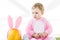 Kid in pink fluffy costume holding colorful quail eggs while sitting near yellow ostrich egg with bunny ears headband