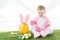 Kid in pink fluffy costume sitting near yellow ostrich egg with bunny ears headband, colorful Easter eggs and tulips