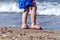 Kid `s Legs on the Sandy Beach by the Sea. Boy with Blue Swimsuit Walking on the Sand During Summer Time. Wave in Background