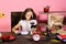 Kid and school supplies on pink background. Girl sits at desk with stationery, clock, books and flowers