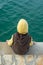 Kid with a yellow hat and brown vest sitting on a stone pier near the green sea water