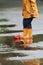 Kid in yellow waterproof cloak and boots playing with paper handmade boat toy outdoors after the rain