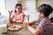 Kids are sitting at table. She is eating cookie. Boy holds cup of milk. They are looking at each other and smiling