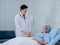 The kind Asian male doctor in white suit visits, talks and gives support, holding hand of happy elderly senior woman patient.