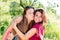 Kissing fun: brunette young women best friends having joyful time laughing & looking at camera on green su