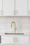 A kitchen faucet detail with a gold faucet, brown backsplash, and white cabinets.