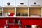 Kitchen interior at home or small office. Red tile wall and white shelf. Cabinet doors with text labels. Glass jars with coffee,