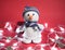 A knitted white snowman with a carrot for a nose sits on a red Christmas background with lights and decorations. Charming snowman