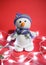 A knitted white snowman with a carrot for a nose sits on a red Christmas background with lights and decorations. Charming snowman