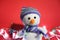 A knitted white snowman with a carrot for a nose sits on a red Christmas background with lights and decorations. Charming snowman