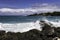 Kona Coast, Hawaii`s Big Island. Wave breaking on rocky shore. Blue-green ocean. Shoreline with trees beyond. Cloudy blue sky.