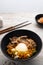 Korean instant noodle with stir fried brisket and onsen egg in black bowl on wooden table
