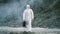 Lab technician in a mask and chemical protective suit, walks on dry ground with a tool box through toxic smoke