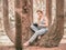 Lady in denim overalls writes something in a notebook while sitting on a multi-stem tree in the park. Post-processing