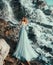 Lady in light long blue flying dress, water nymph stands near large waterfall